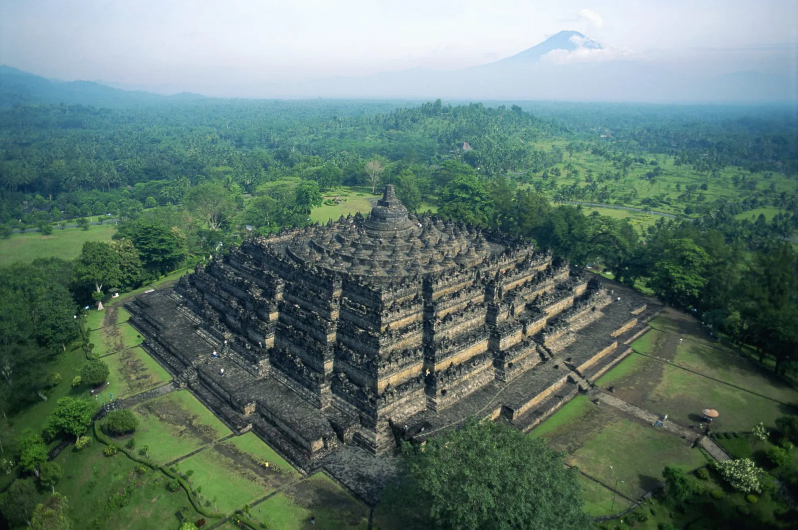 Borobudur from above, its square terraces among the trees, a volcano on the horizon