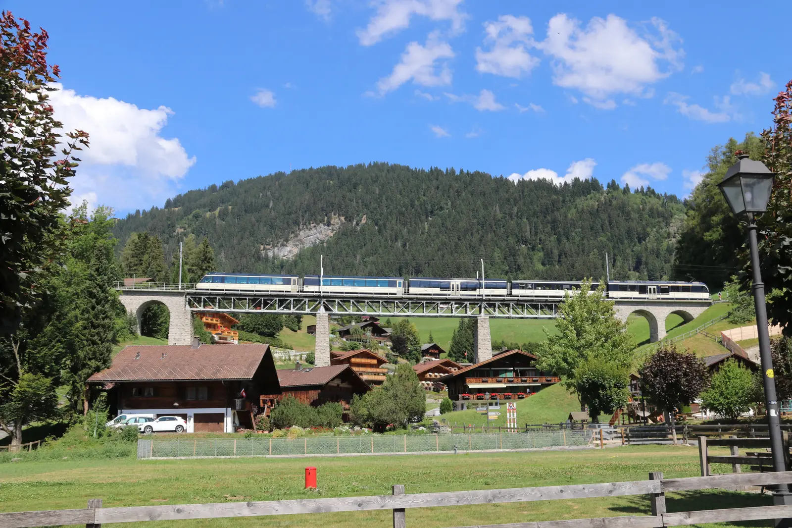 The GoldenPass panoramic train crossing the Grubenbach viaduct above the chalets of Gstaad, with a wooded hill behind and a meadow in front