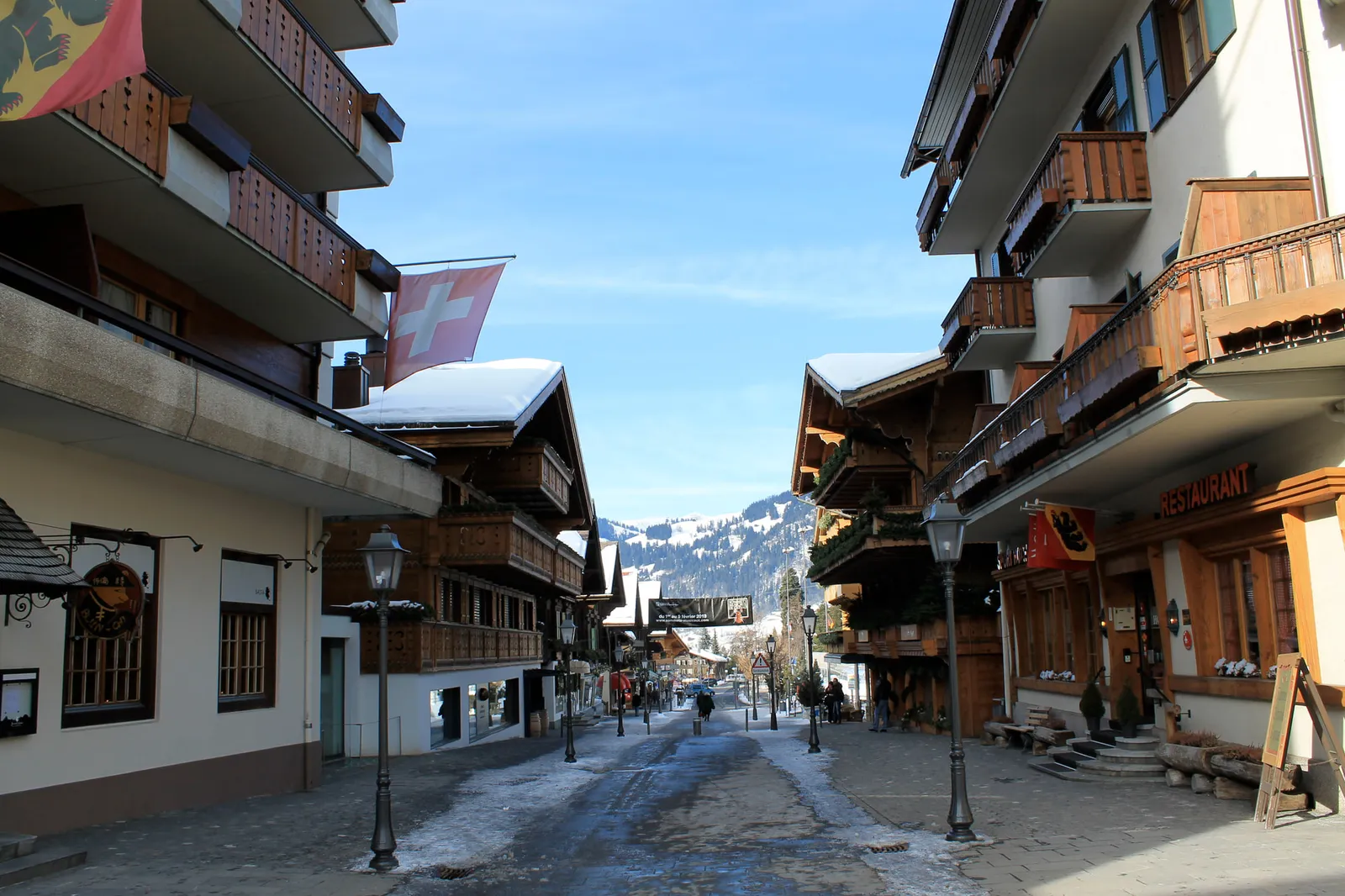 The Promenade in winter: chalets and wooden balconies, street lamps, Swiss and Bernese flags, snow along the edges and ski slopes at the end of the street
