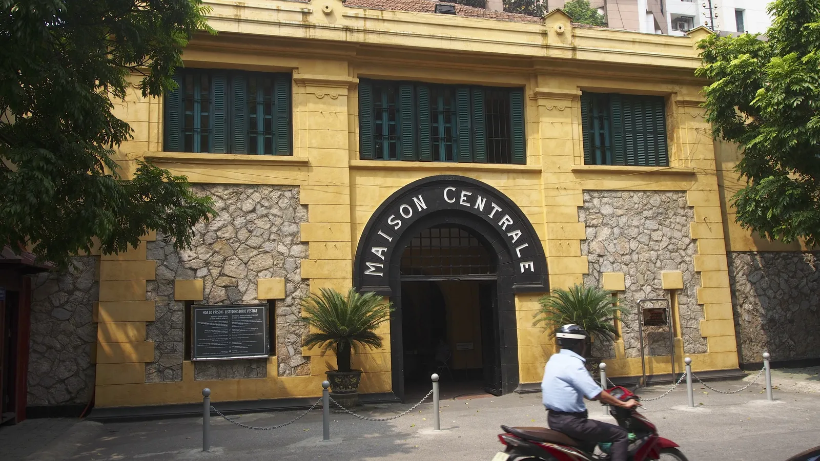 The yellow entrance of Hoa Lo Prison under its «Maison Centrale» arch, a moped in front