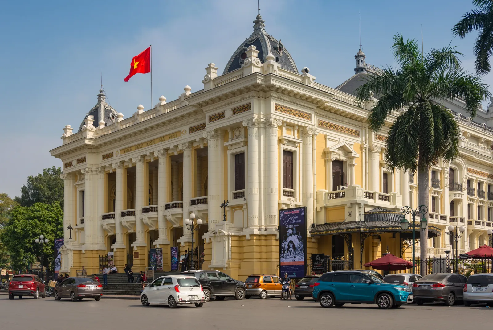 The yellow and white façade of the Hanoi Opera House with its domes and a palm tree, cars in front