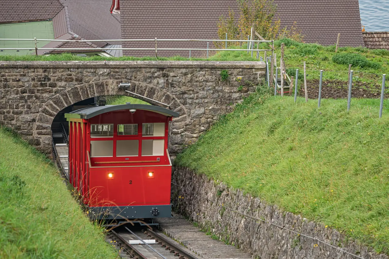 A red car of the Bürgenstock funicular coming out of a stone arch below Kehrsiten, between grassy banks