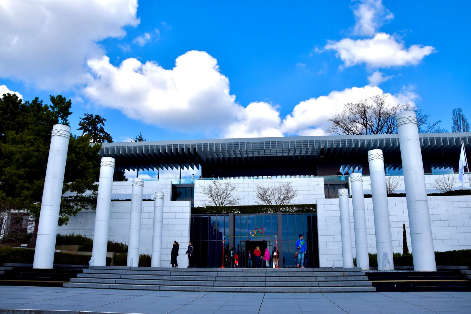 The entrance to the Olympic Museum: white columns before the white façade, broad steps and the Olympic rings above the glass doors, under a blue sky with clouds