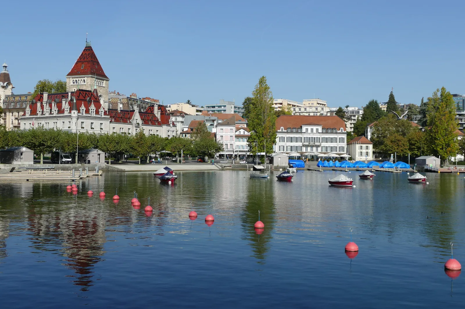 The Château d’Ouchy and its red-roofed tower seen from the Vieux-Port: moored boats, red buoys on calm water and trees along the lakefront