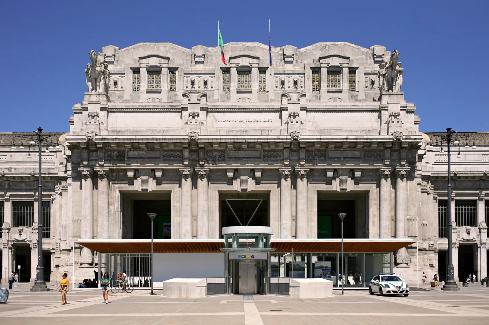 The facade of Milano Centrale seen head-on from Piazza Duca d’Aosta, under a blue sky
