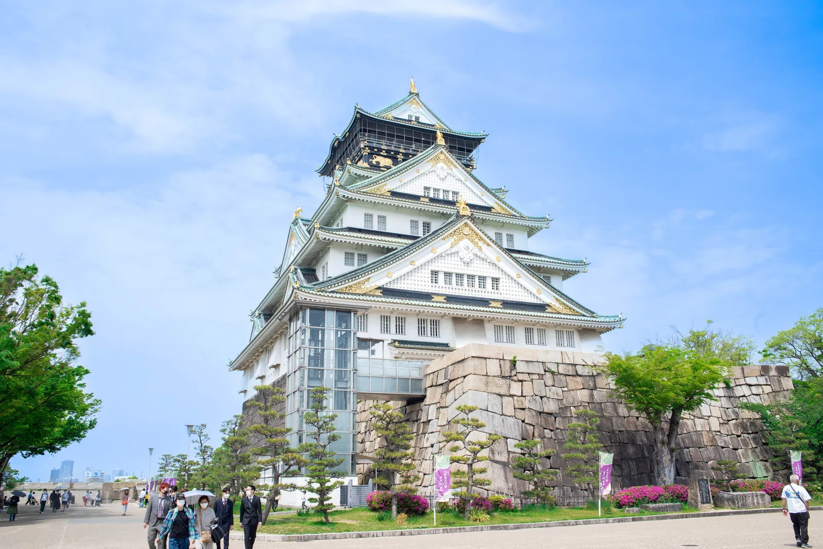 The white and green main tower of Osaka Castle on its stone base, visitors on the path below