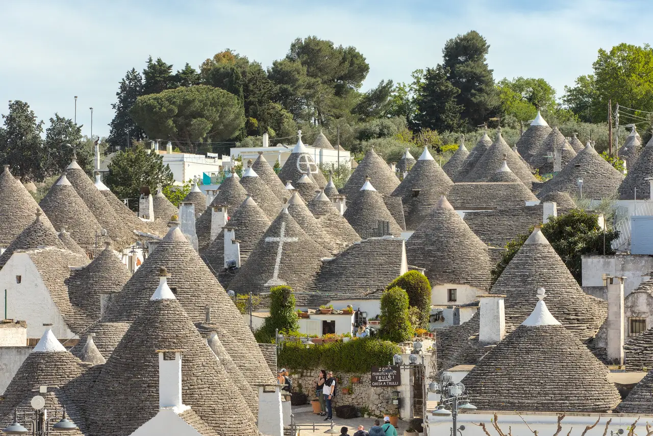 The trulli of Rione Monti in Alberobello: rows of conical stone roofs, some painted with white symbols, and trees on the hill behind.
