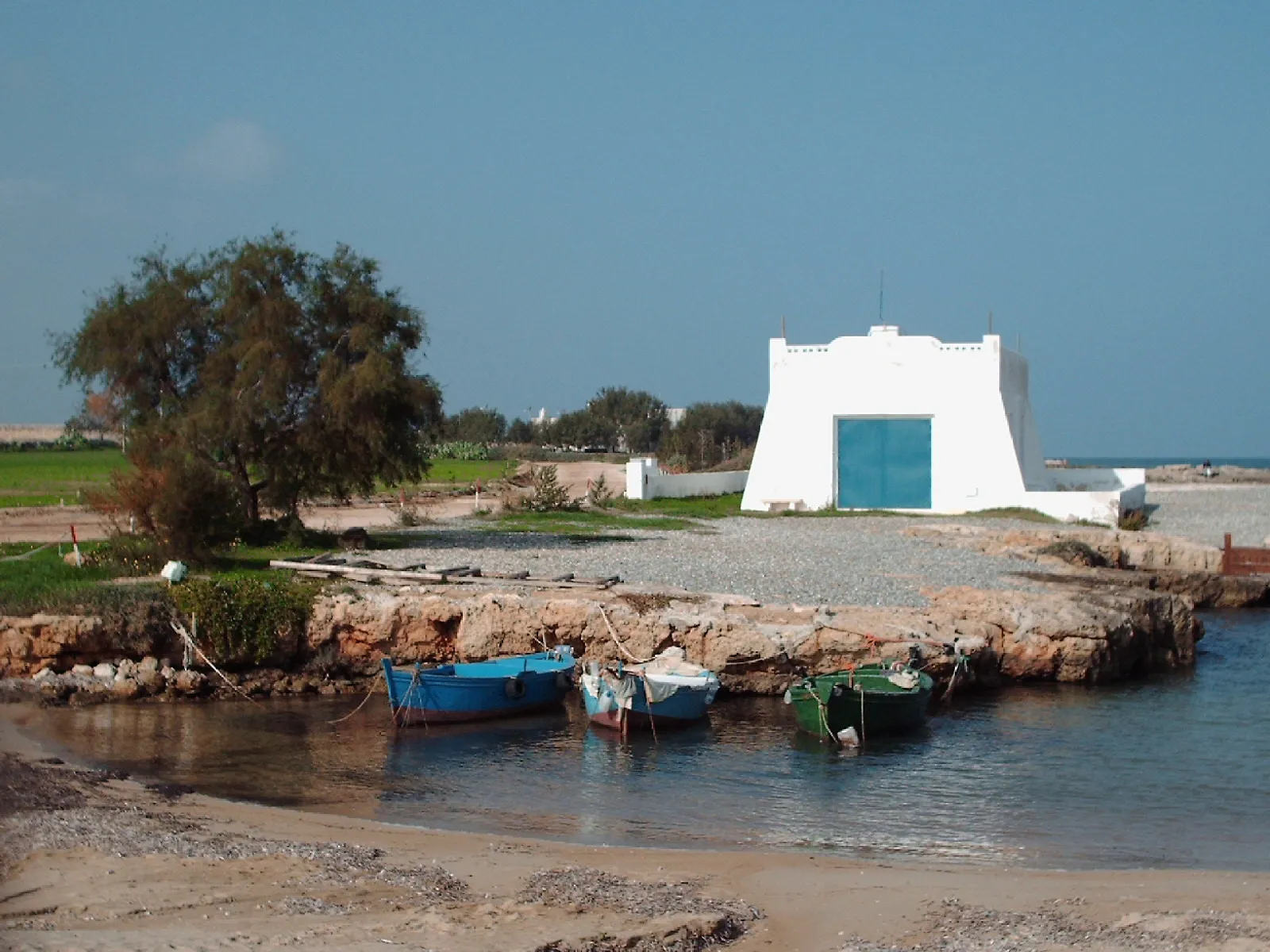 3 small fishing boats moored in a rocky cove on the Savelletri shore, in front of a whitewashed building with a blue door.