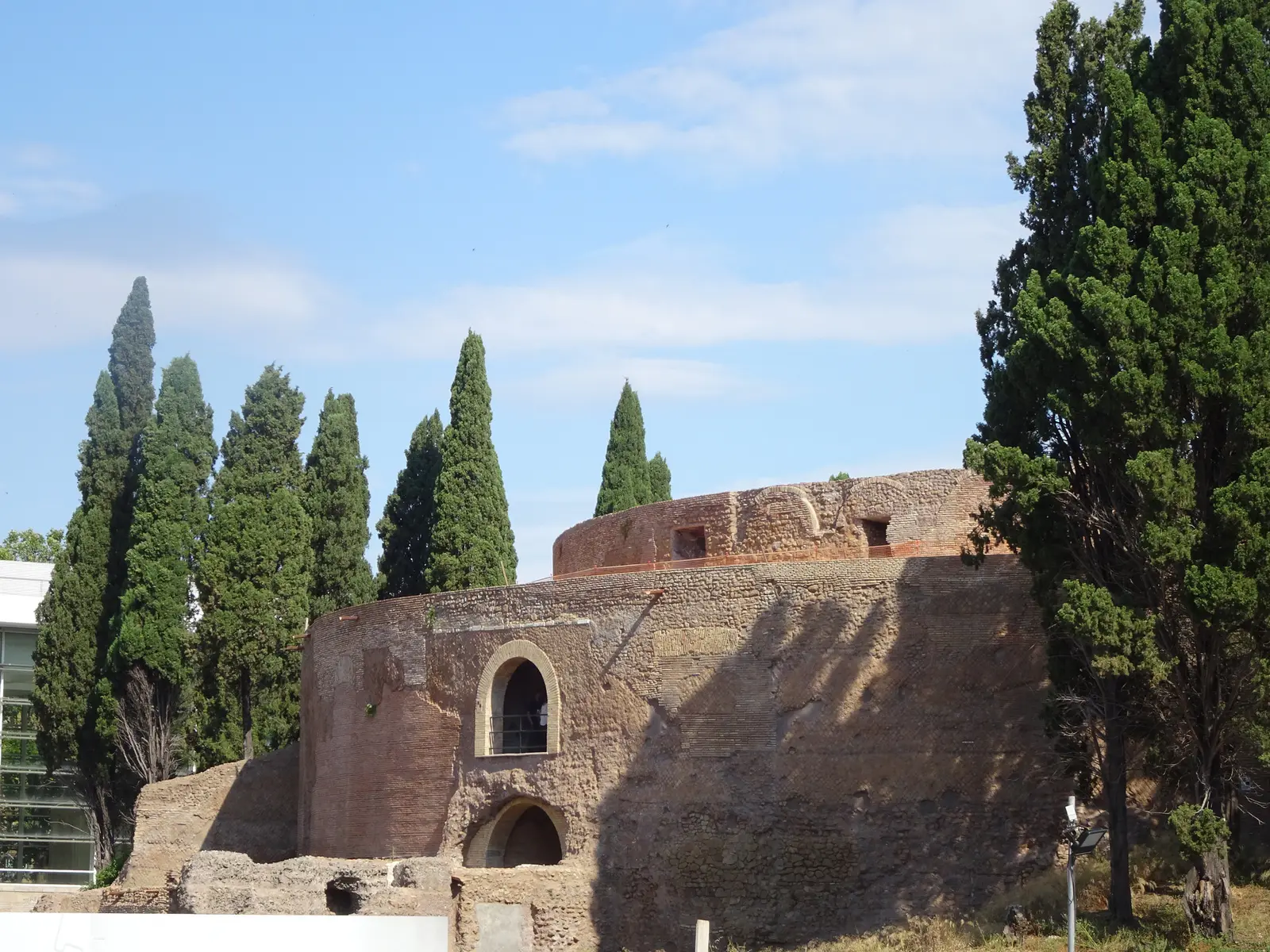 The Mausoleum of Augustus after its restoration: the round brick drum with its arched openings among cypresses, under a blue sky.