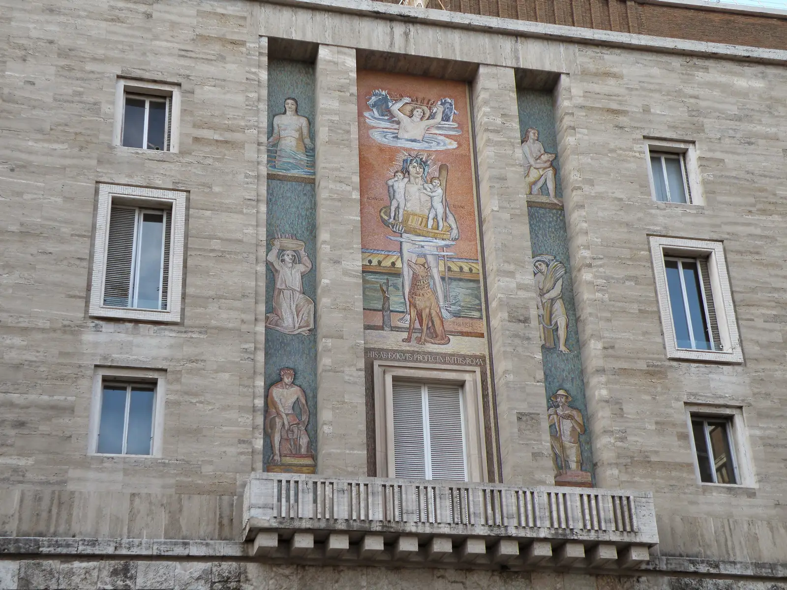 The mosaic on the building’s façade: the Tiber, standing in the river, holds the basket with Romulus and Remus, the she-wolf at his feet; in the side panels, women and labourers on a green ground