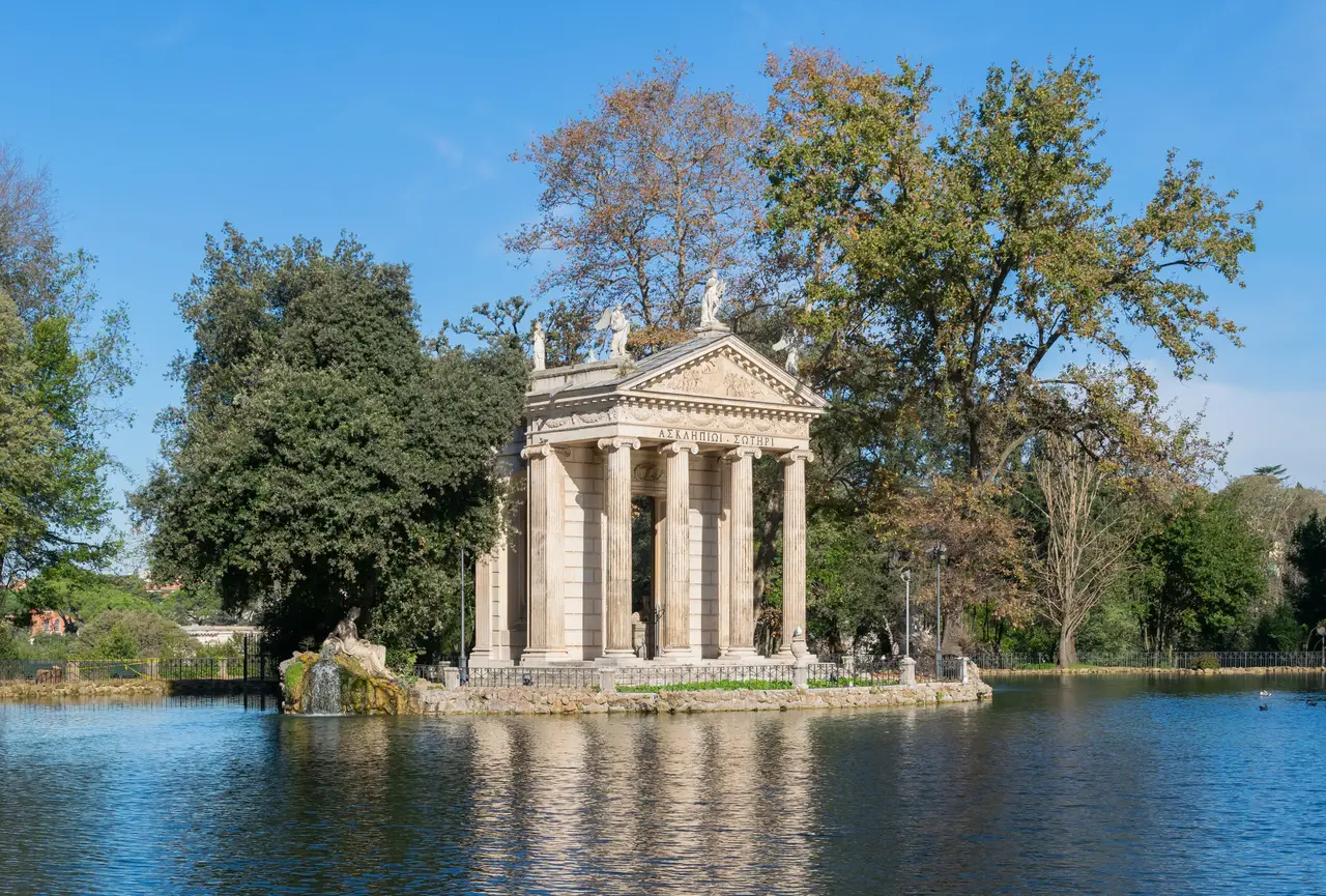 The lake at Villa Borghese with the Temple of Aesculapius on its island: Ionic columns and statues on the pediment reflected in the water, among trees beginning to turn