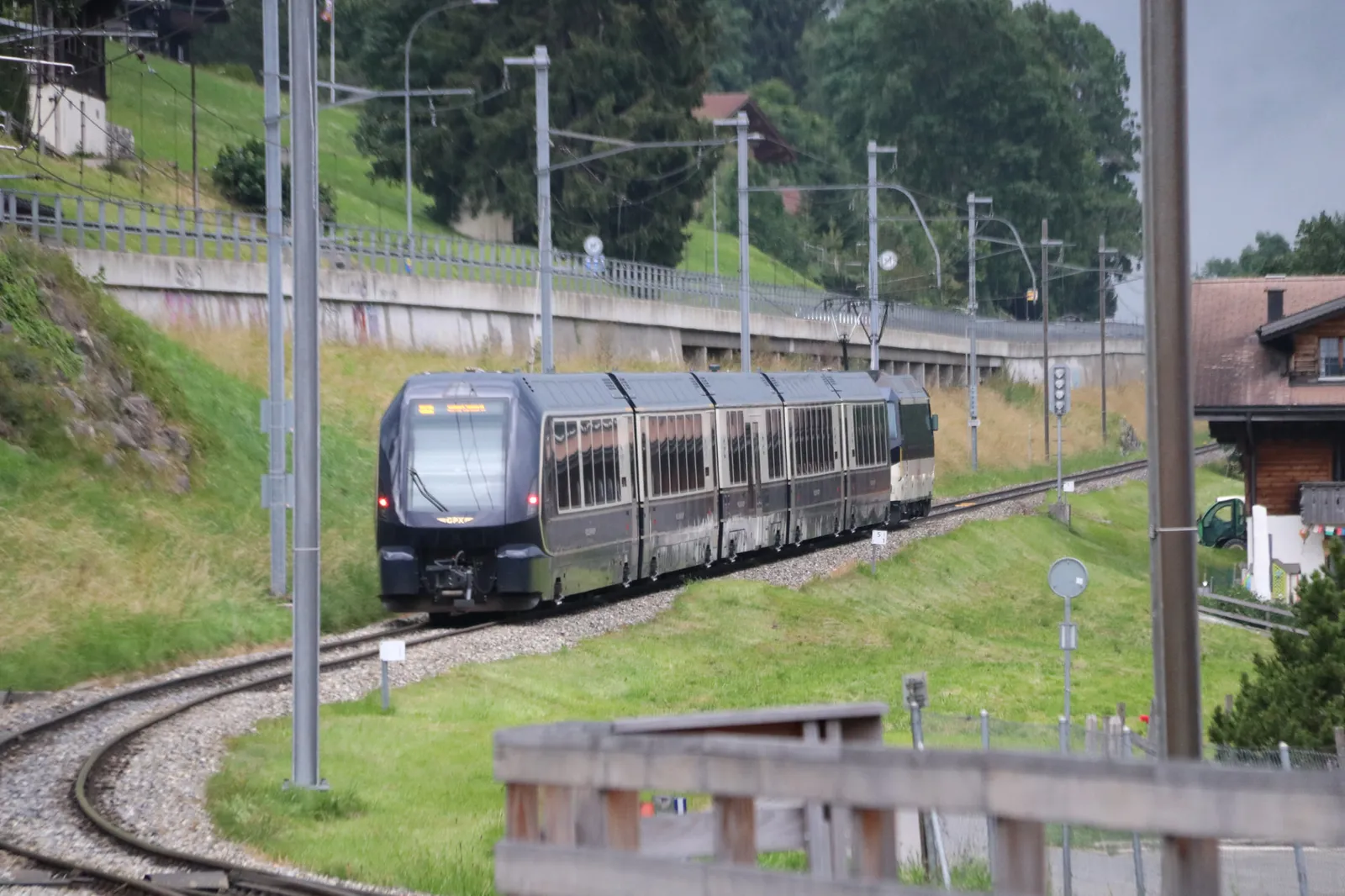 The GoldenPass Express on a curve of the line near Schönried, between overhead-line masts, a green slope and a chalet