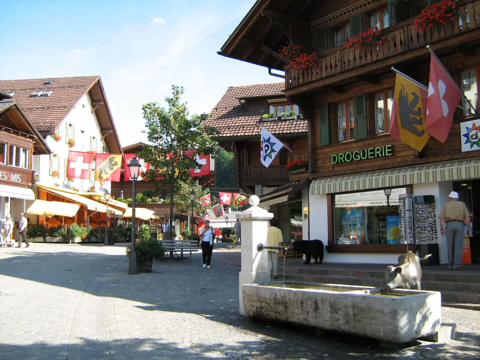Gstaad’s main street in summer: chalets with red geraniums and Swiss and Bernese flags, a drugstore, a stone fountain and people walking
