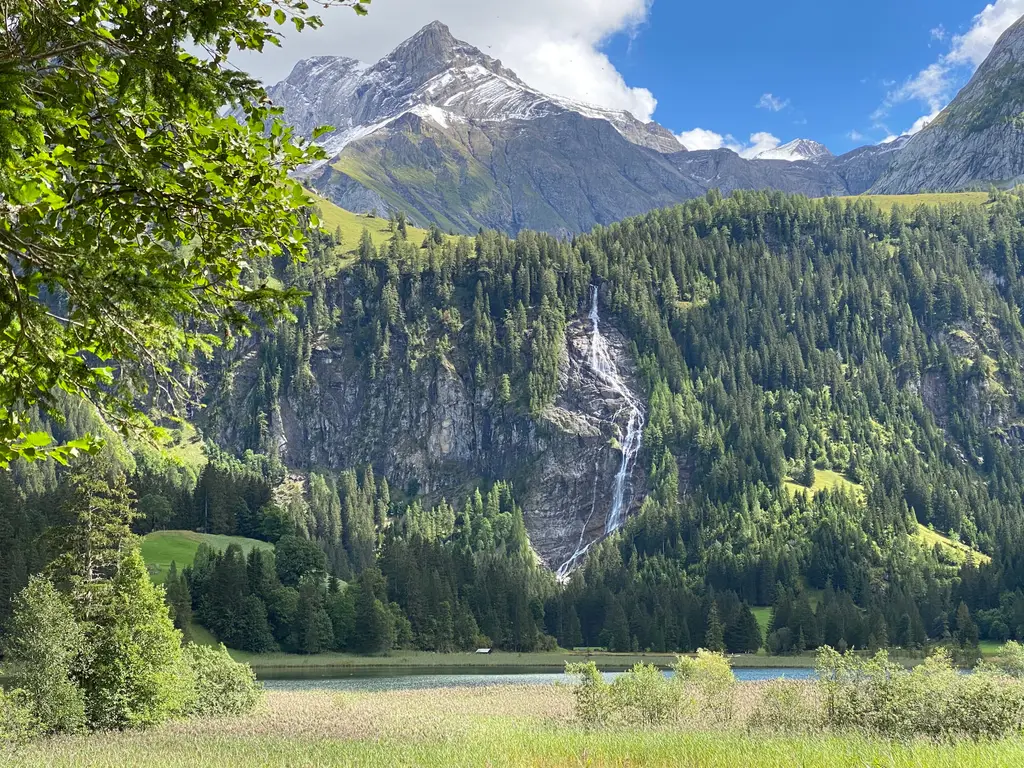 At the Lauenensee: reeds and a strip of the lake, the Tungelschuss waterfall on a rock face, forested slopes and a snow-streaked peak