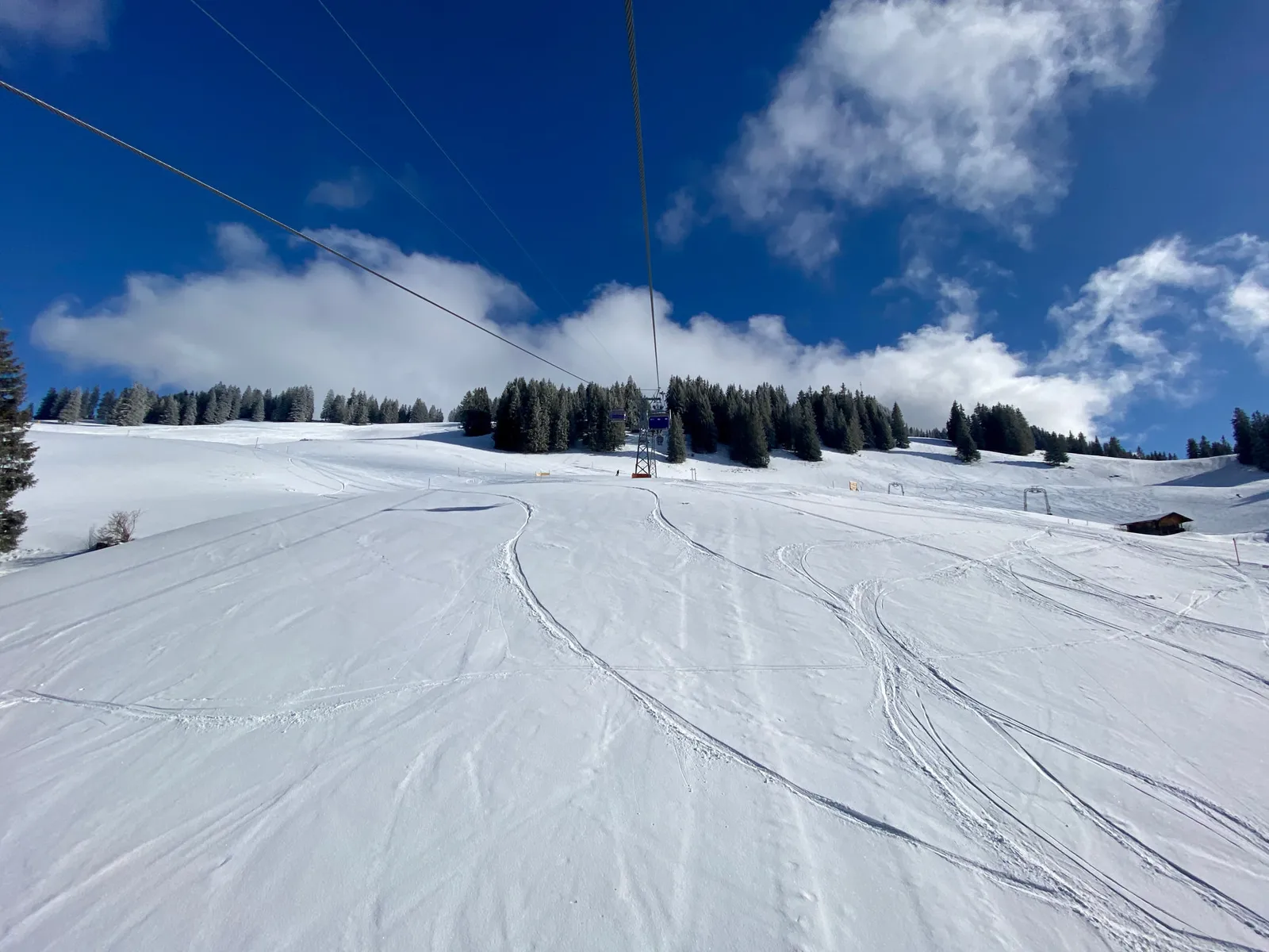 A ski slope on the Horneggli above Schönried: fresh tracks in the snow under a chairlift cable, fir trees, a wooden hut and a blue sky with clouds