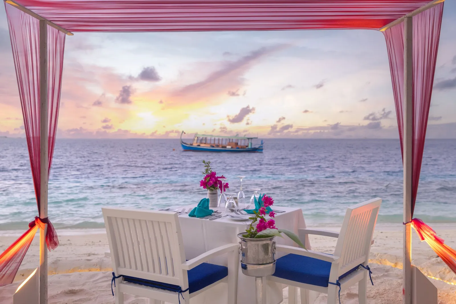 A table for 2 under a canopy on the beach, a dhoni offshore at sunset