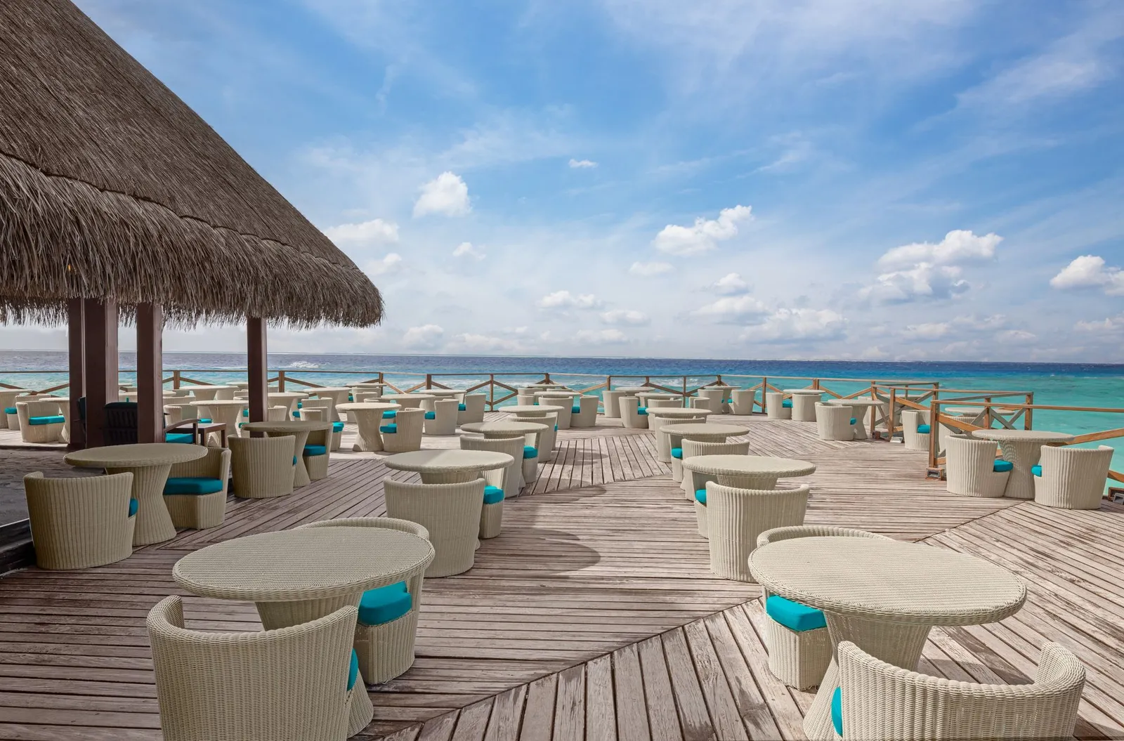 A bar’s wooden deck over the sea, white tables and wicker chairs under a thatched roof