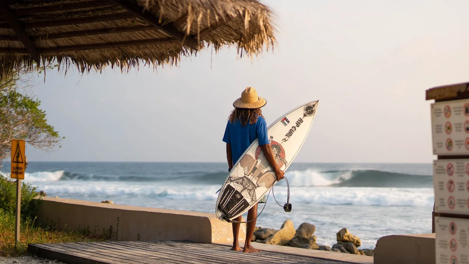 Someone with a surfboard watches the waves from the beach deck