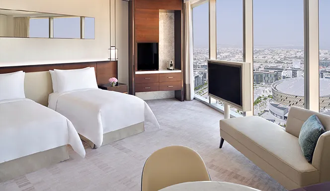 A room with 2 beds side by side and the city through the glass
