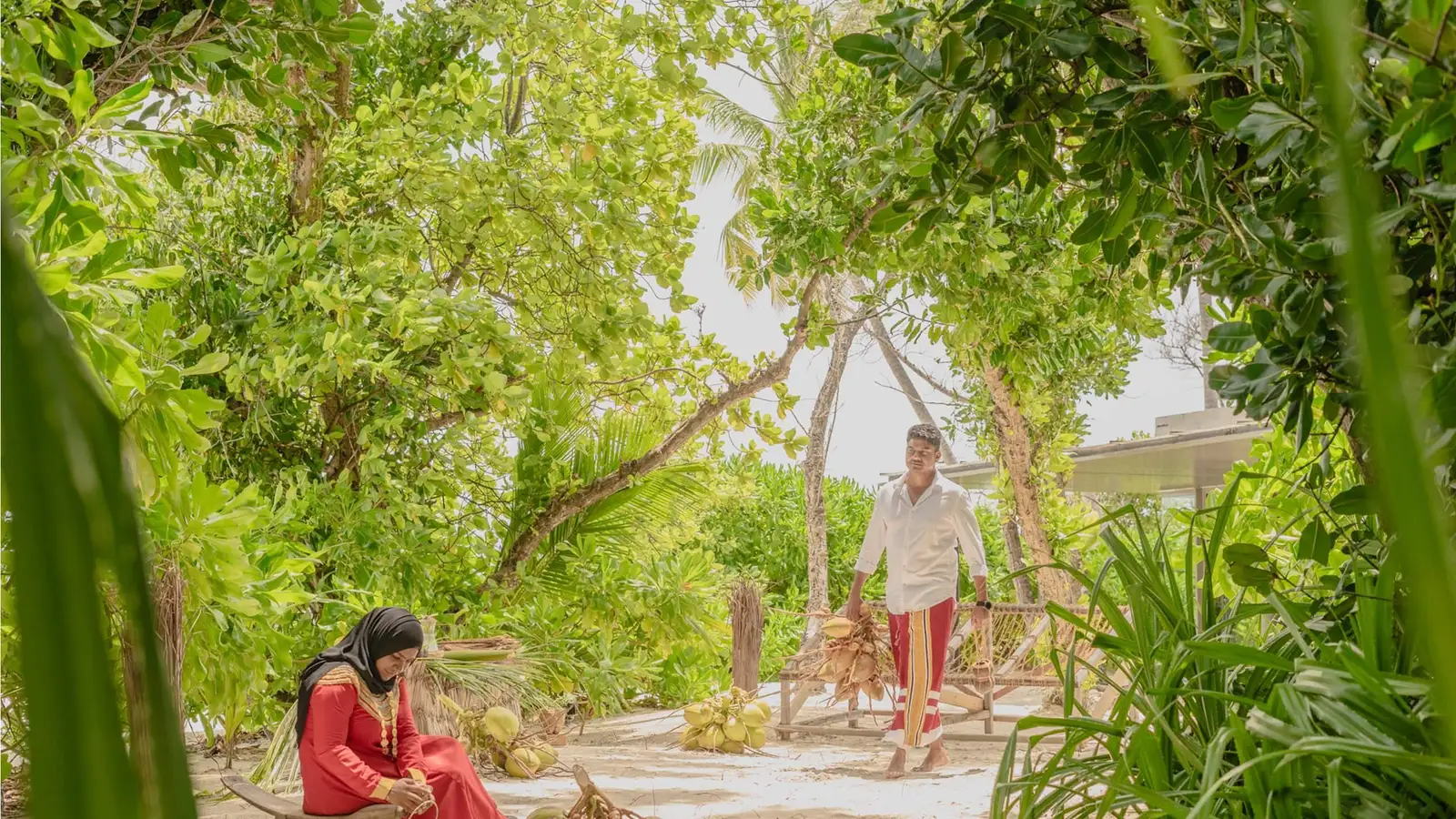 2 people working with coconut and leaves under a shaded shelter.