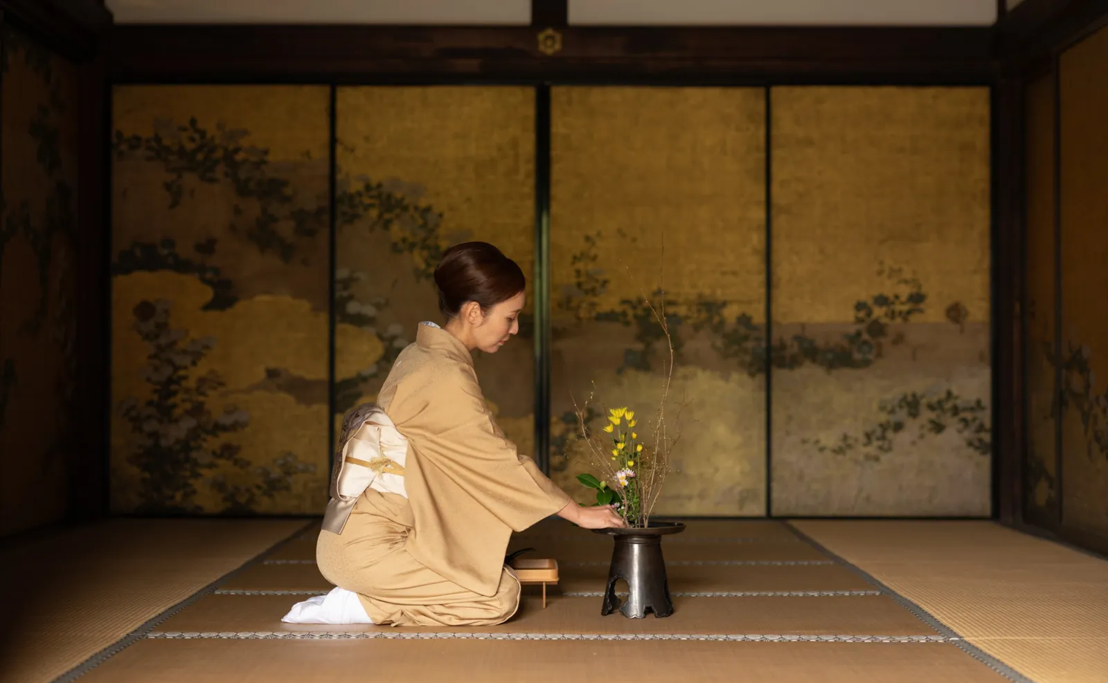 A woman in a pale kimono, kneeling on tatami before a gold screen, arranges yellow flowers in a dark vessel.