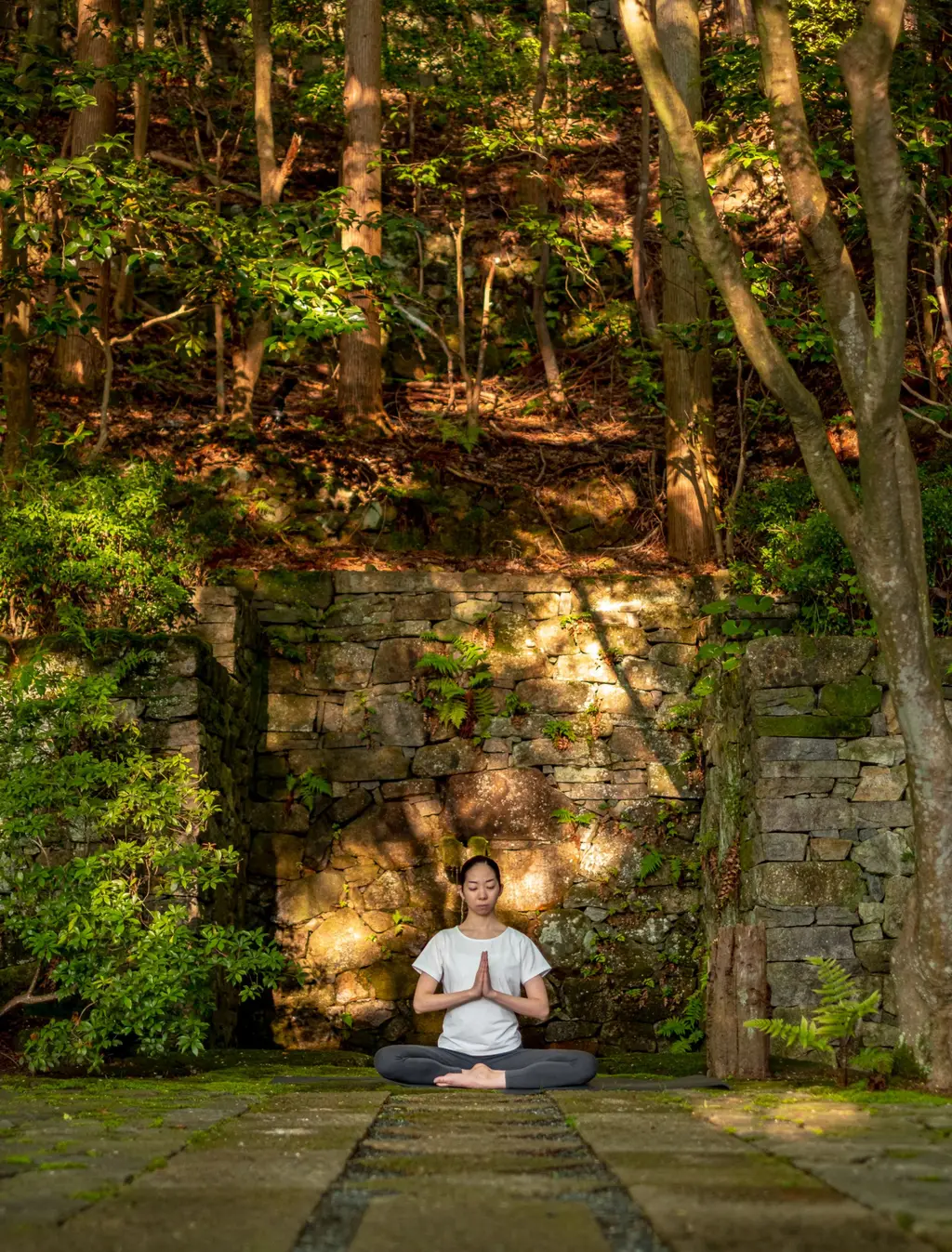 A man sitting cross-legged on the stone path, at the foot of an old stone wall among the trees.