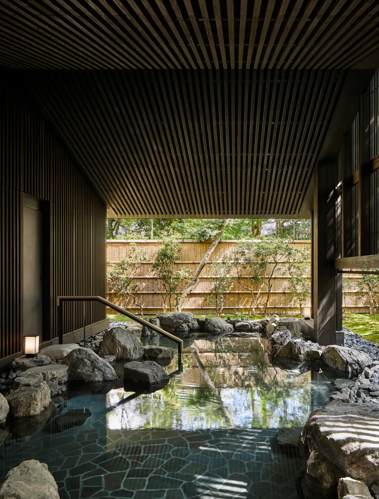 The outdoor onsen pool among rocks, under the slatted timber ceiling, with the bamboo fence and the green beyond the edge.