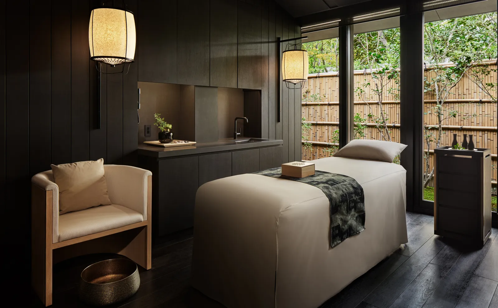 A spa treatment room with its bed, an armchair, paper lanterns and the glass wall onto the bamboo fence.