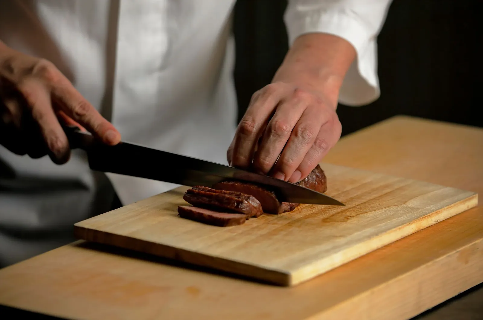 A cook’s hands slice a piece of cooked beef on a pale wooden board.