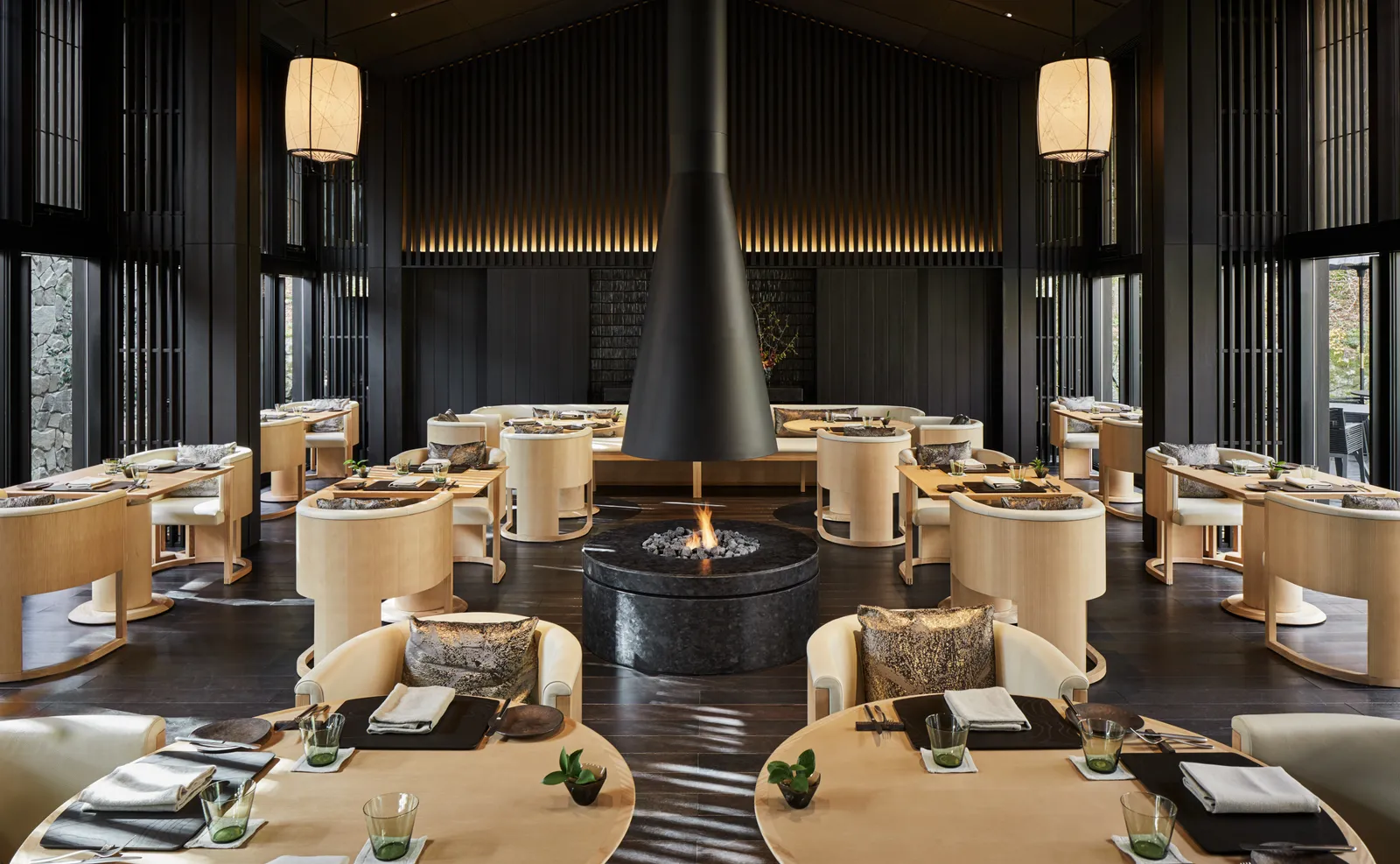 The Living Pavilion’s room, the black cone fireplace in the middle, pale armchairs, paper lanterns and windows onto the forest.