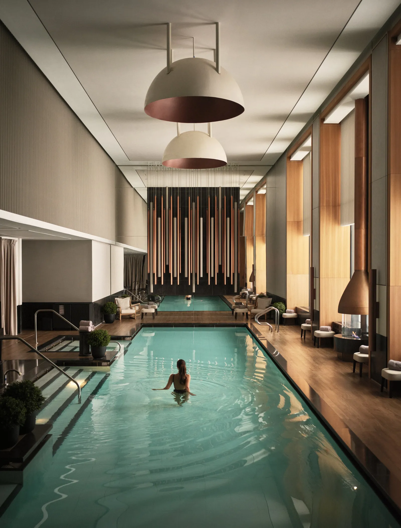 Aman New York’s indoor pool beneath 2 large lamps, with timber columns and warm light