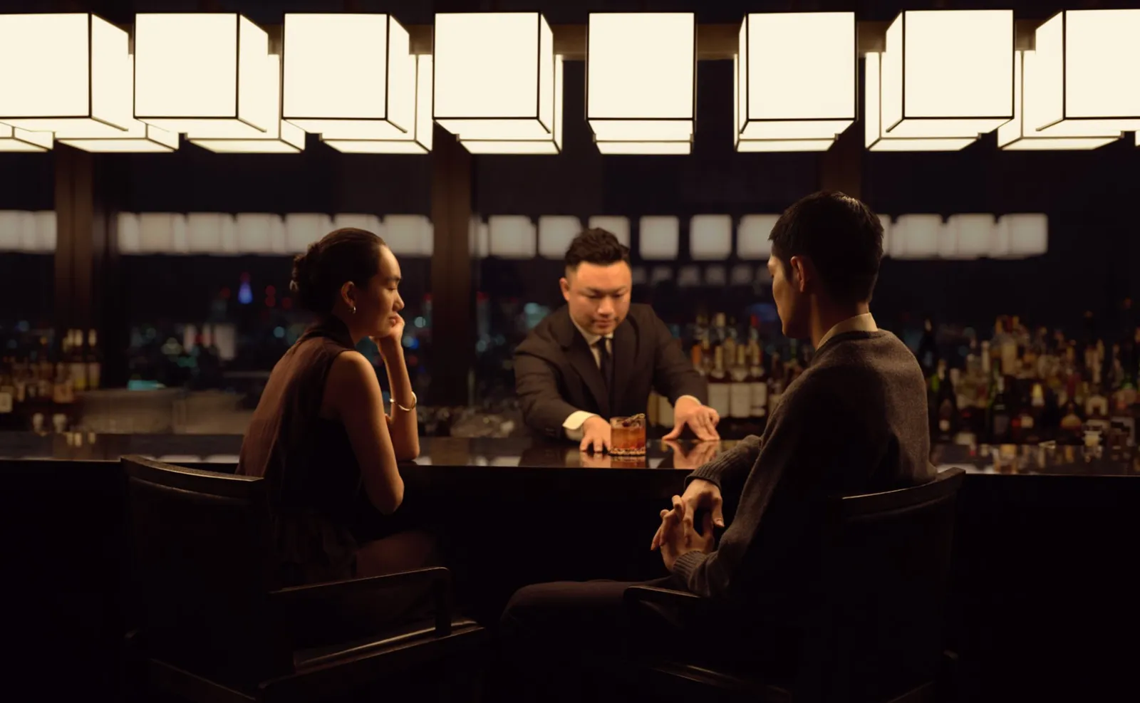 The Lounge’s bar counter at night, under white lanterns: a bartender serves 2 seated guests.