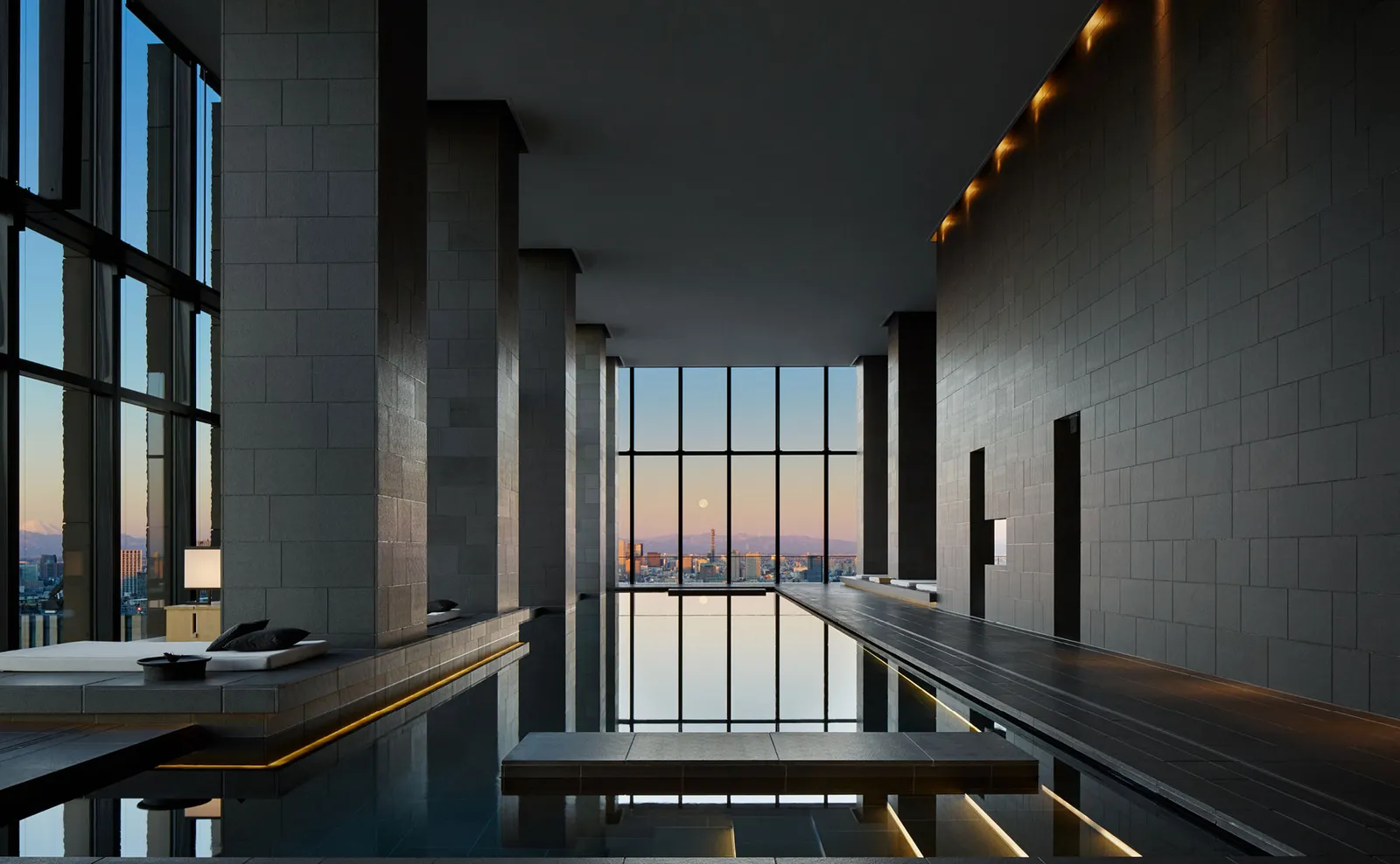 The spa pool at dusk, long between dark stone walls, with daybeds and the window onto the city at the far end.