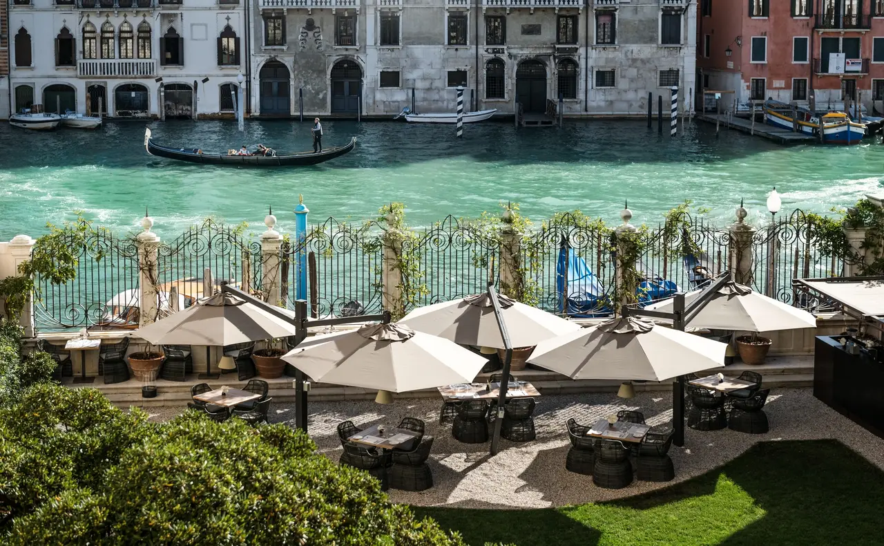 Arva’s tables under parasols in the garden, along the railing on the Grand Canal, with a gondola and the palazzi opposite.