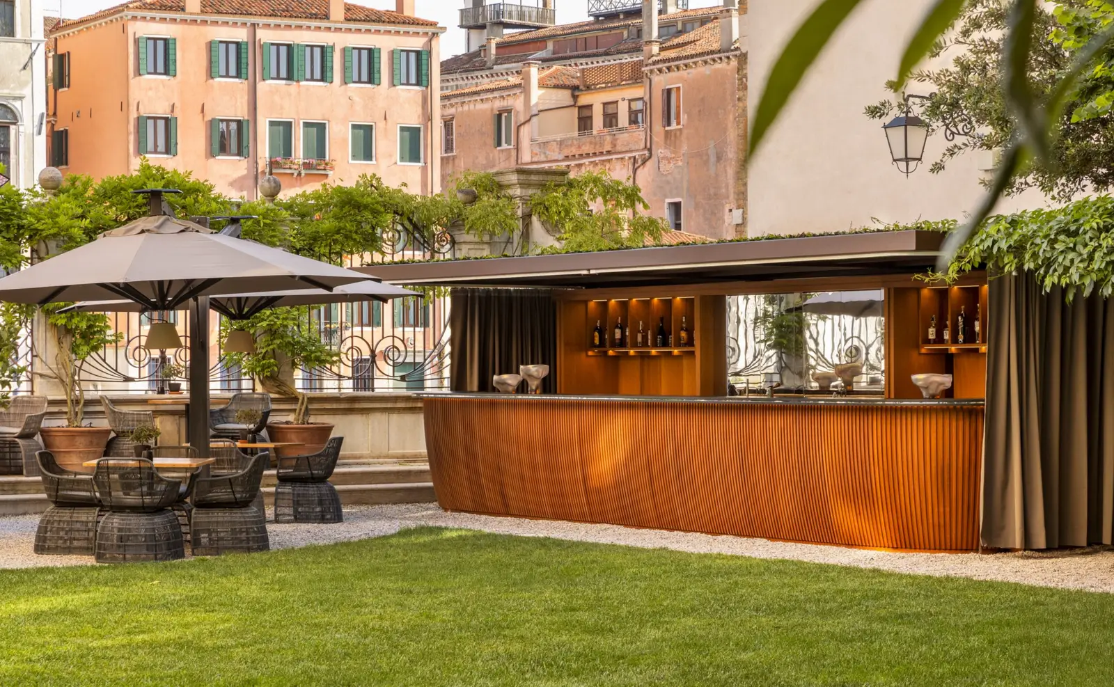 The outdoor bar in the garden: the fluted timber counter, parasols and armchairs on the lawn, the palazzi on the Grand Canal behind.