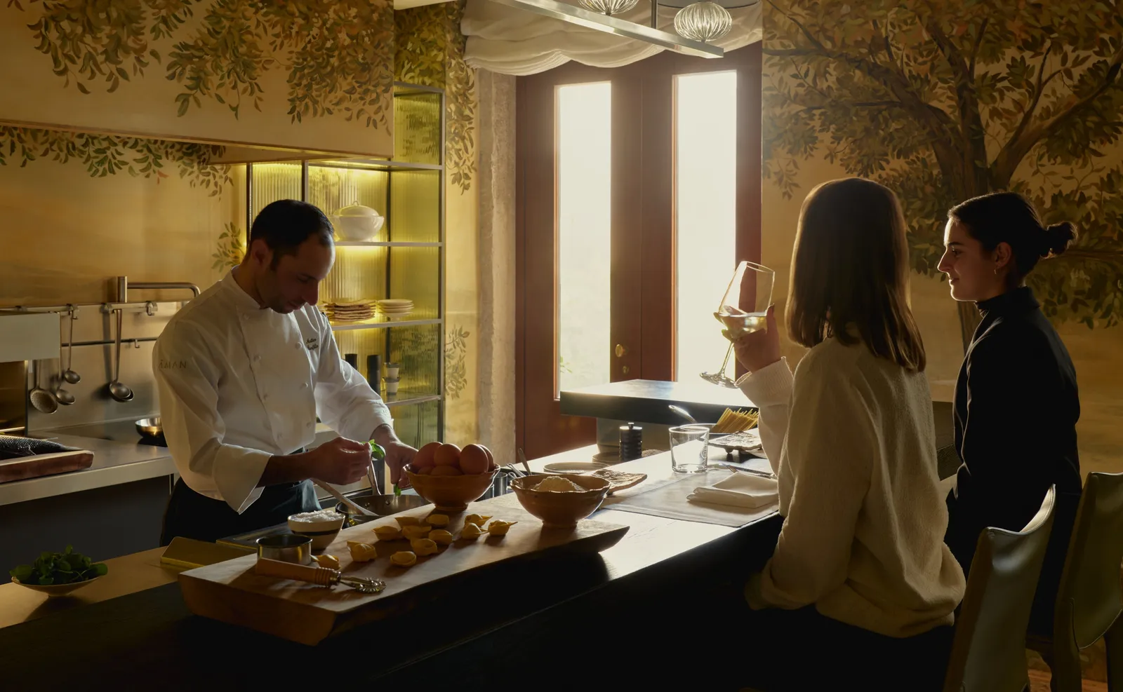 At the Palazzo Kitchen Table a chef prepares dishes at the counter while 2 guests, glasses in hand, watch him work.
