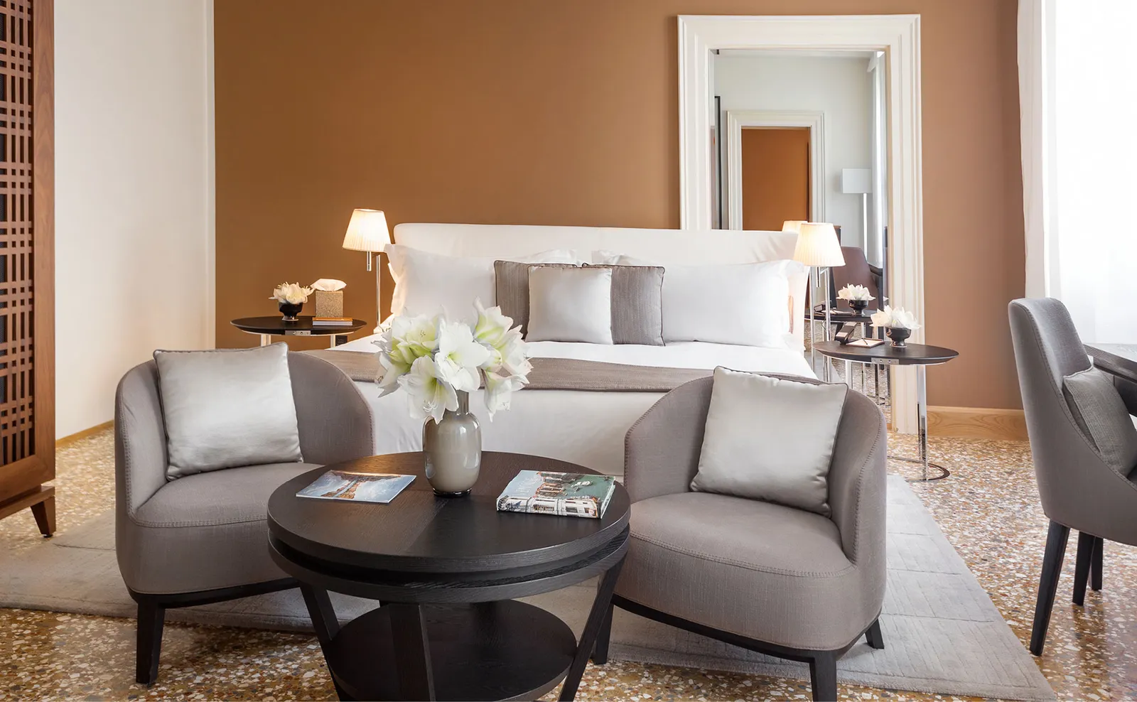 The sitting area of a Palazzo Chamber Luminoso: 2 grey armchairs, a round table and, behind, the bed against the ochre wall.