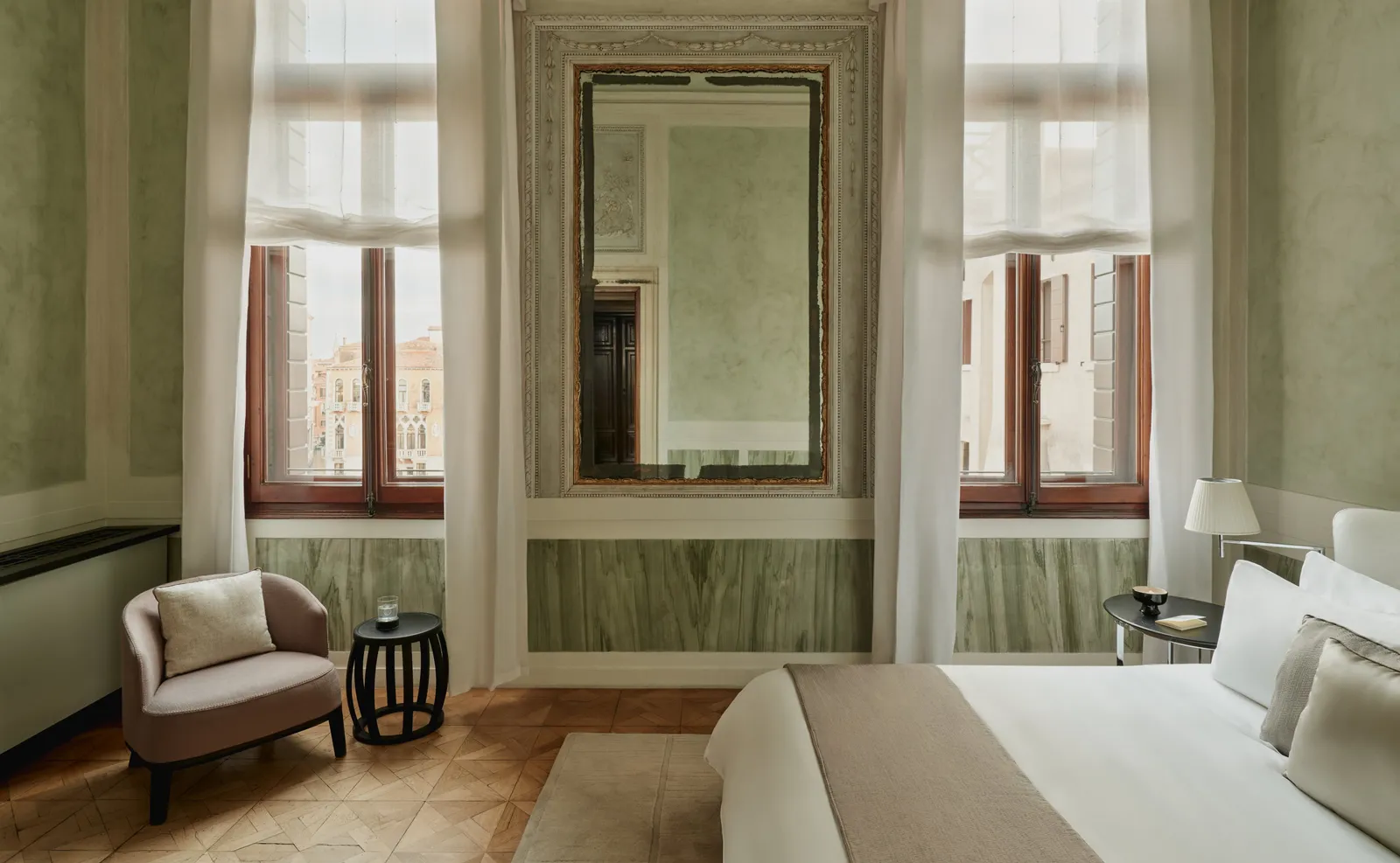 A Palazzo Stanza Canal Grande: sage-green walls, an old mirror between 2 windows and the bed in the foreground.