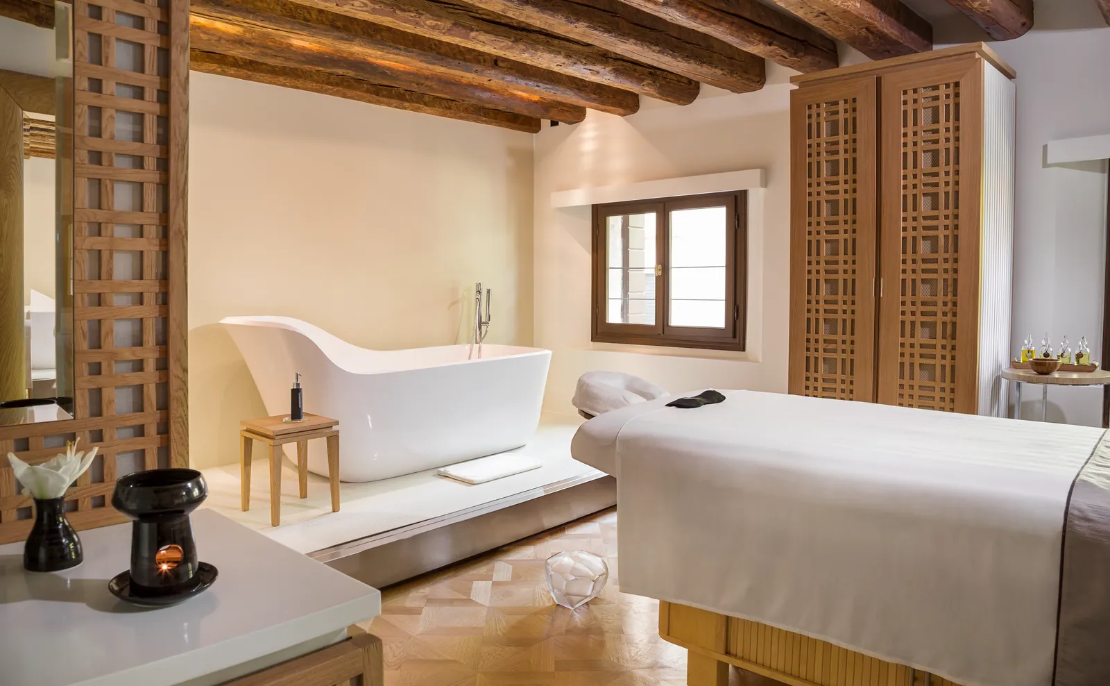 A spa treatment room under timber beams: the freestanding tub, the massage bed and a window onto the rooftops.
