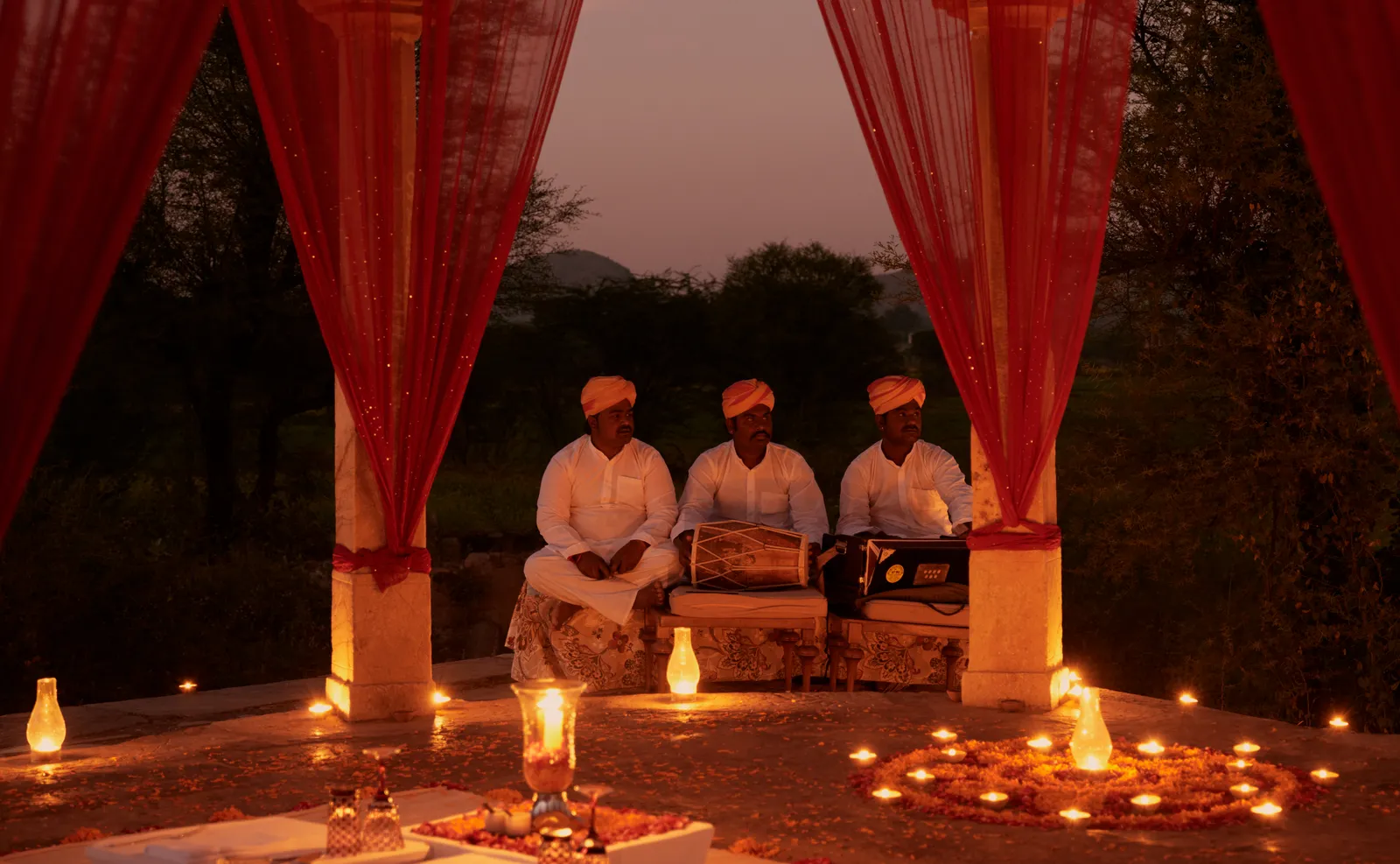 Musicians seated beneath the red drapes of a chhatri, among lit clay lamps