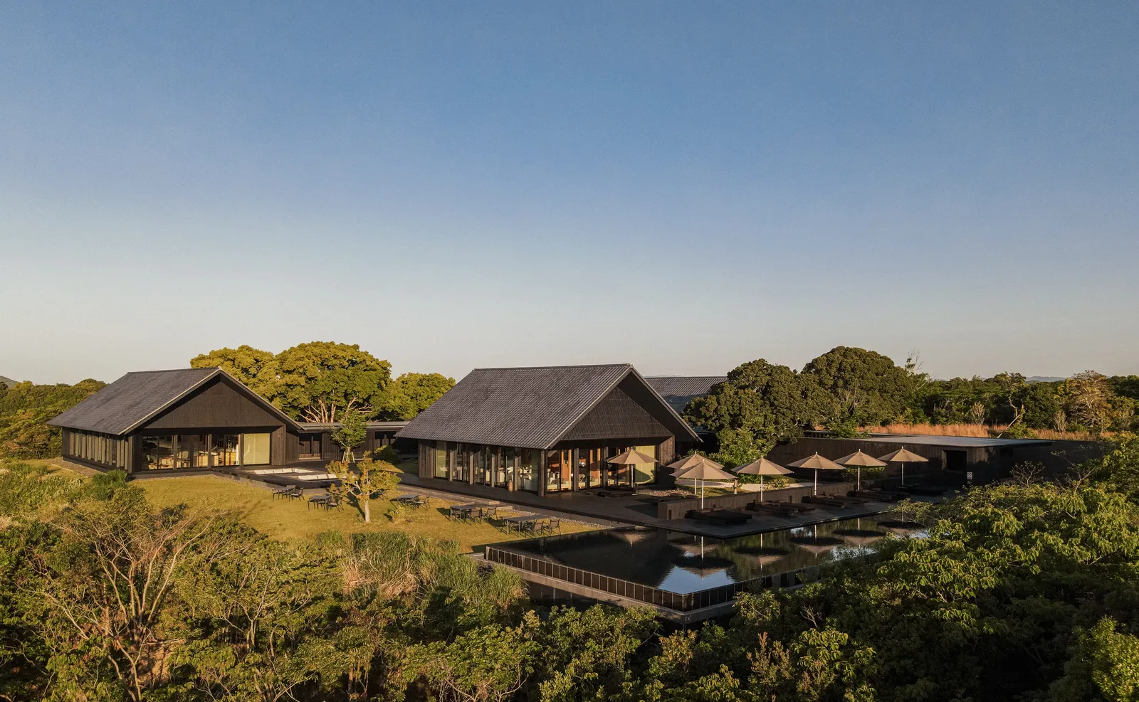 2 dark-roofed pavilions and the pool terrace on the forested hillside, the bay behind