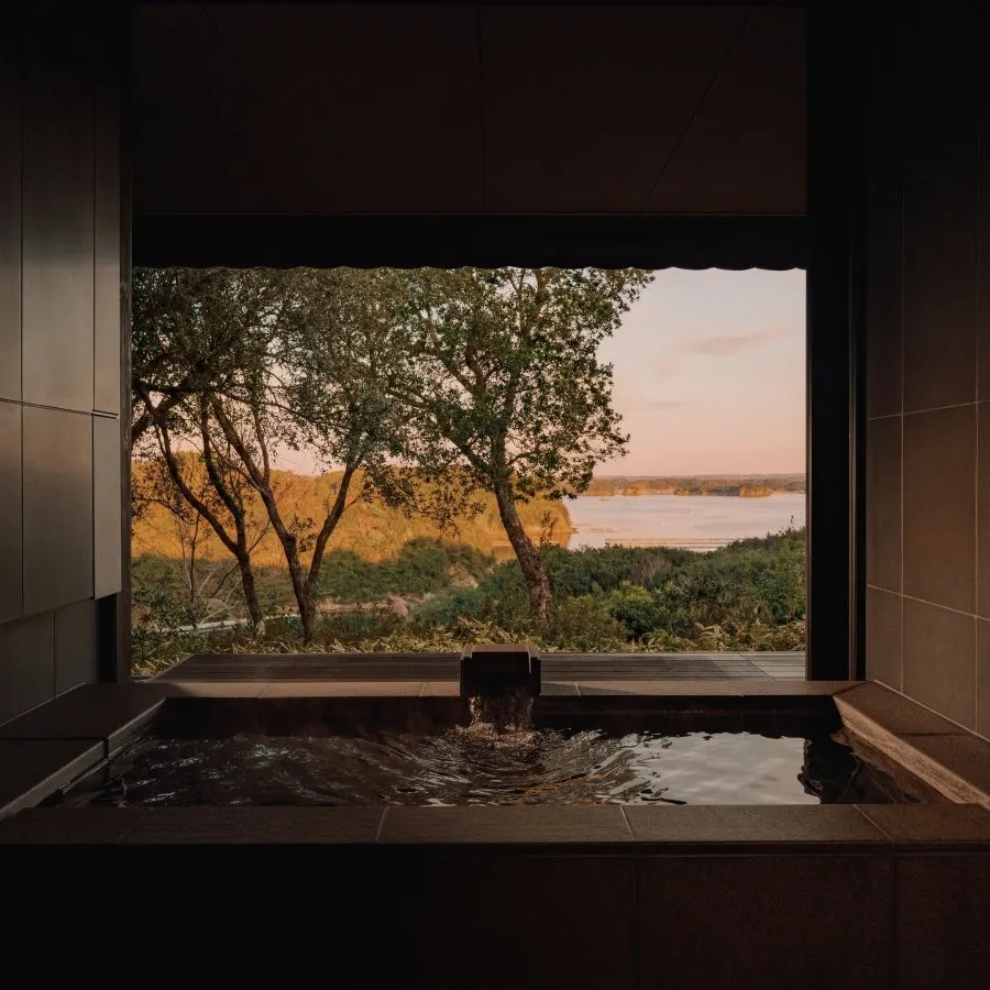 A stone onsen at a window over trees and the bay at sunset