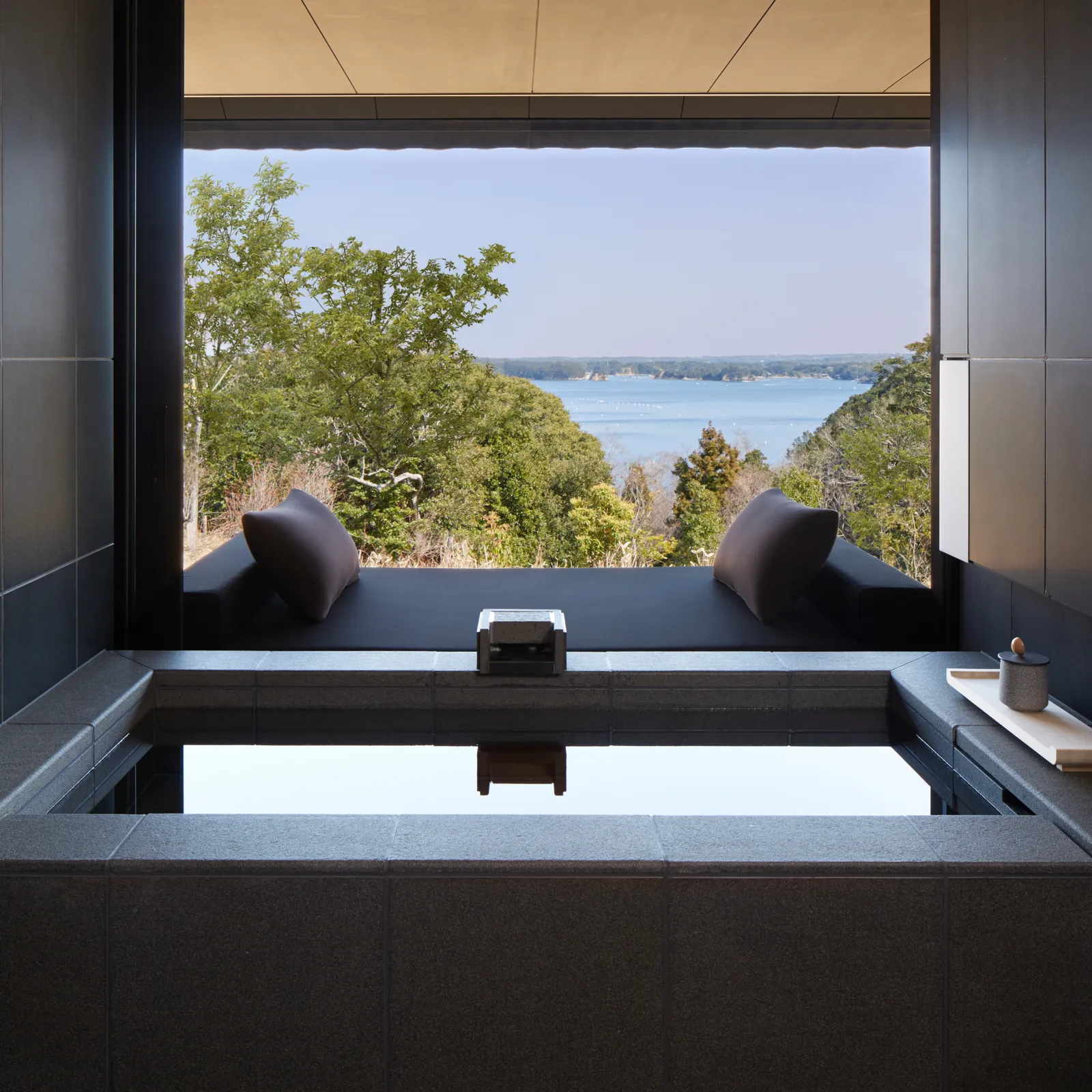 A suite’s stone hot-spring bath at an open window, the bay beyond the trees