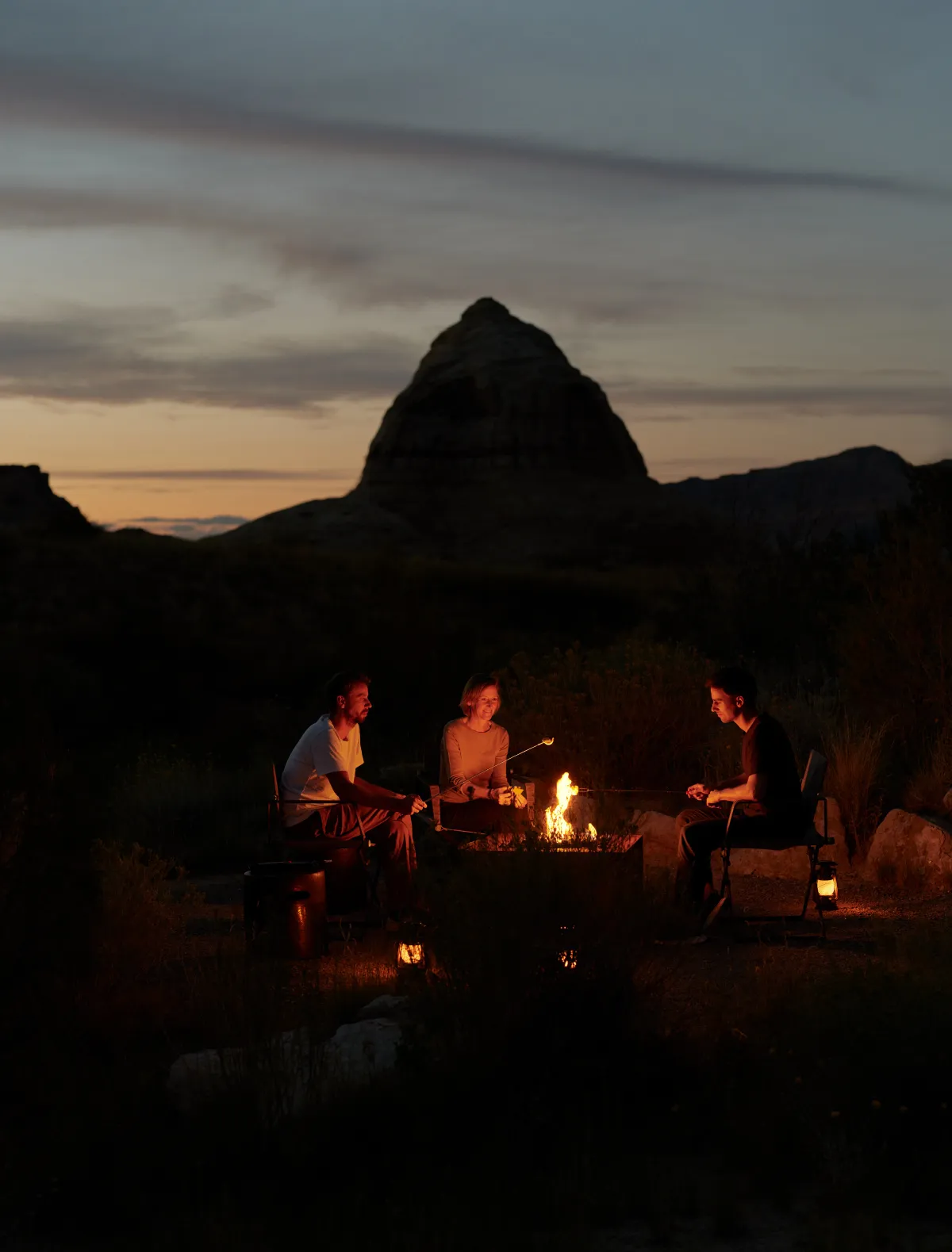 3 people sitting round a fire at nightfall, beneath the dark outline of a butte.