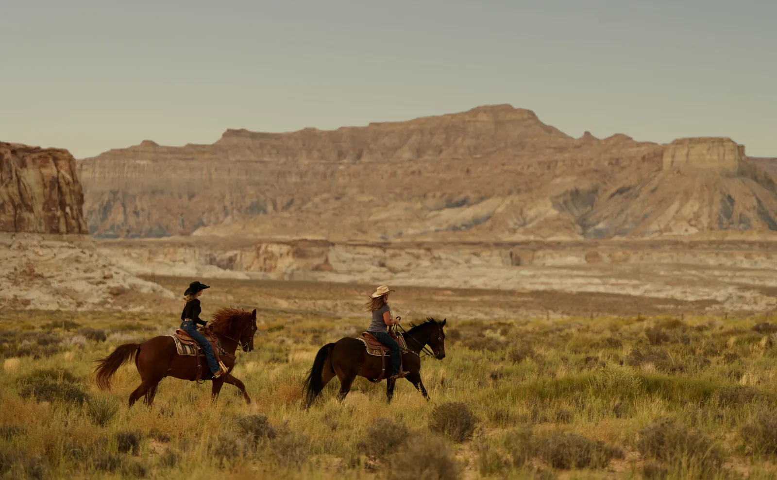 2 riders cross the scrub plain on horseback, a long mesa behind them.