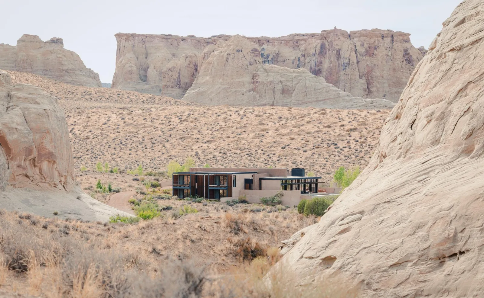 The Mesa Home from a distance: a low house with dark windows between 2 rock walls, beneath a mesa.
