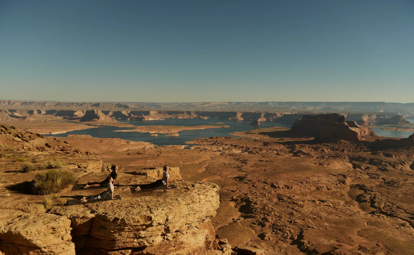 2 people practise yoga on a rock ledge, above the canyons and waters of Lake Powell to the horizon.