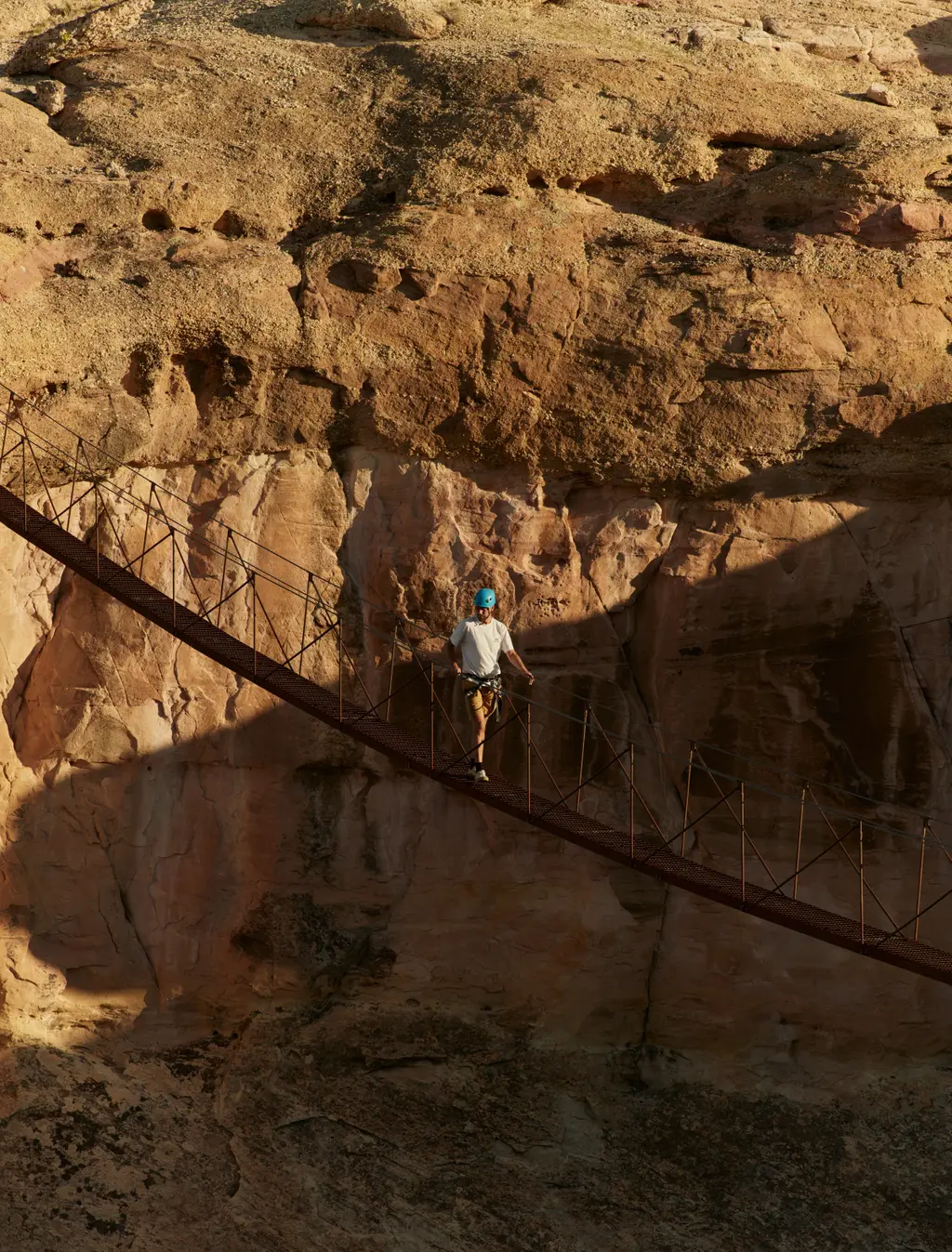 A person in a helmet crosses a suspension bridge of cables and steps before a sandstone wall.