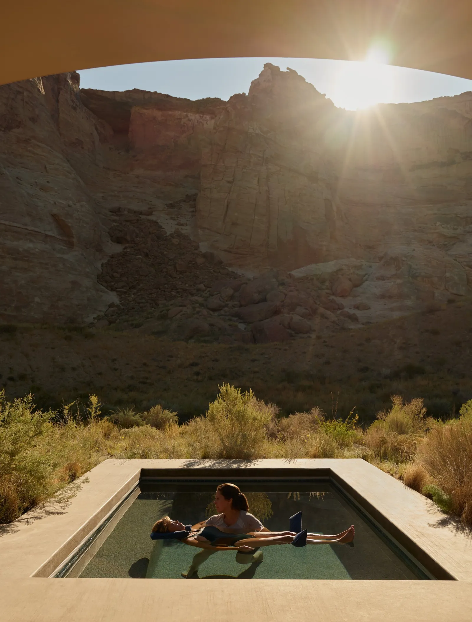 A therapist supports a guest floating in a small open-air pool, the sun above the cliff.