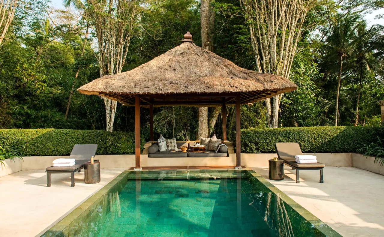 A green-stone pool in front of a thatched balé, loungers on either side